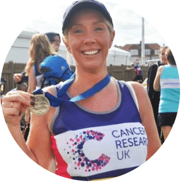 Photograph of Bridie, wearing a baseball cap and Cancer Research UK running vest, holding up her medal. 