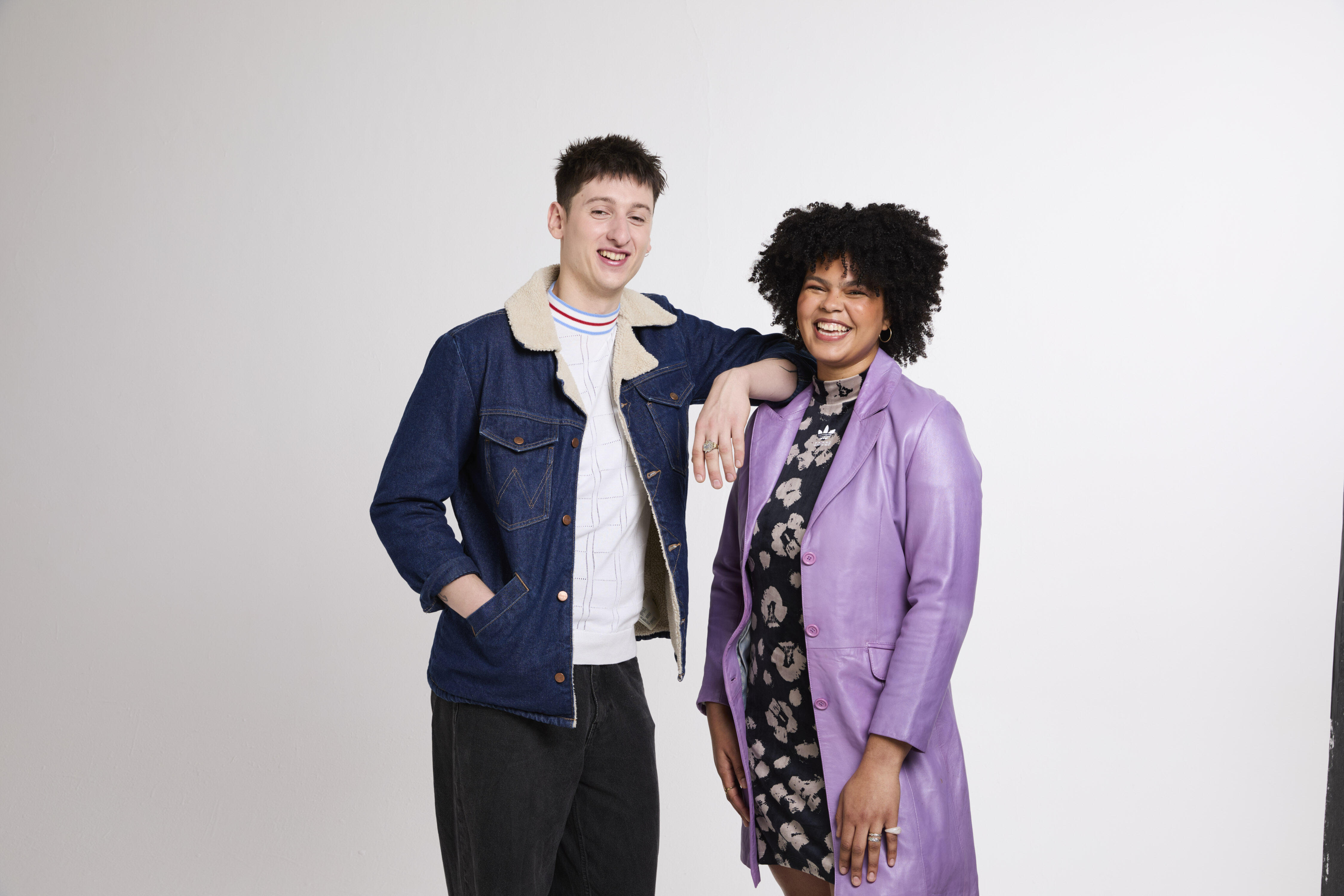Two models smiling, one model is wearing a denim jacket and the other model is wearing a purple coat. 