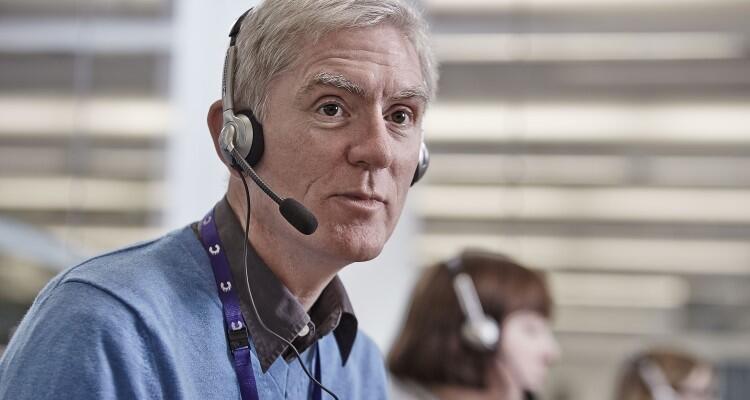 A man wearing a headset, speaking on a call. He appears to be working in an office as you can see others behind him.