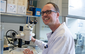 a male researcher wearing a lab coat, smiling in a laboratory next to a microscope
