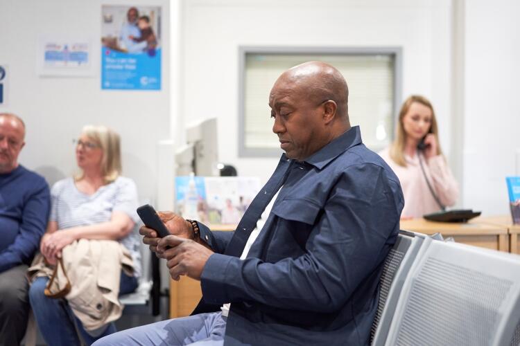 A man sat in a waiting room, looking at his phone. It appears to be a clinical setting.