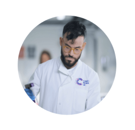 A male researcher in a lab wearing a white coat with the CRUK logo on it and glasses