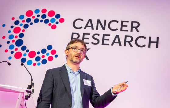 Man stands next to a mic stand talking with Cancer Research UK logo on the wall behind
