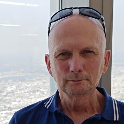 A person with sunglasses perched on top of their head, wearing a blue collared shirt. They are standing in front of a window in a tall building.