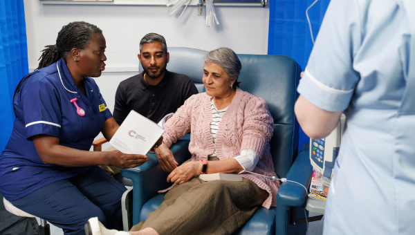 Patient is sat with a family member and a health professional while having chemo. A nurse shows them a Cancer Research UK leaflet.