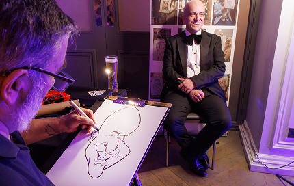 image of man in a suit, smiling, sat on a chair with an artist drawing a caricature portrait of him