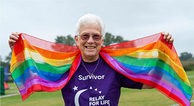 A person holding the Pride flag wearing a purple t-shirt that reads Survivor 