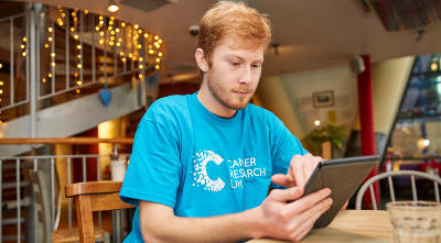 Male e-campaigner wearing CRUK tshirt, sitting in a cafe, using his tablet to e-campaign.