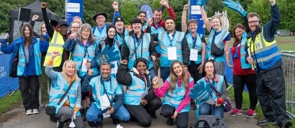 Large group of about 20 volunteers huddled together and smiling to the camera. Gathered by the finish line at the end of an event course.